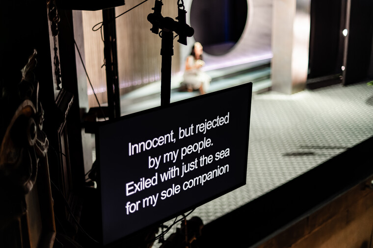 White text on a black surtitle screen displayed in front of a stage