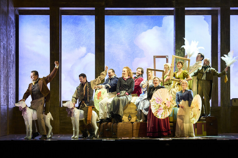 Men and women in period dress form the shape of a carriage on stage in front of a cloud backdrop.