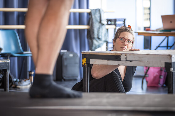 A women sits on the ground fearfully looking up at a man standing on a table
