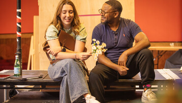 A man and woman sit on benches smiling at each other in rehearsal