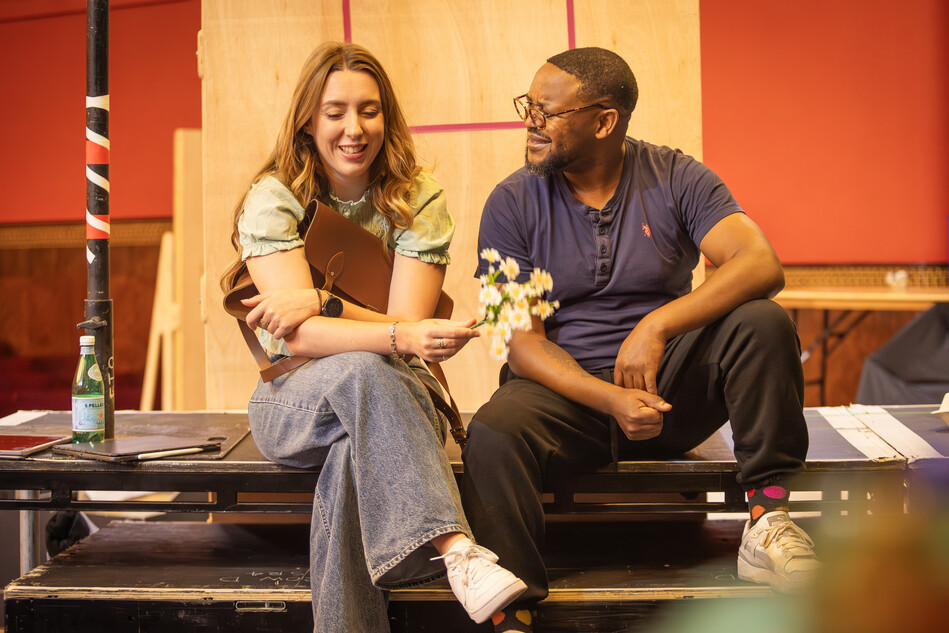 A man and woman sit on benches smiling at each other in rehearsal