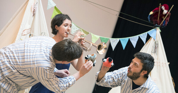 There is a woman playing the trumpet in front of two teepees with bunting in between them. In front of her, two men hold little furry birds up the trumpet like the birds are listening to the music.