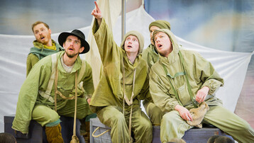 Five men are on a small boat. They are all wearing light green clothes for a cold climate. The man in the middle is pointing up and far away. He lloks concerned by what he sees. The other men are looking in the same direction to see what he is pointing at.