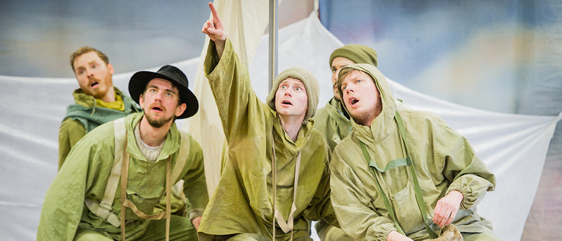 Five men are on a small boat. They are all wearing light green clothes for a cold climate. The man in the middle is pointing up and far away. He lloks concerned by what he sees. The other men are looking in the same direction to see what he is pointing at.