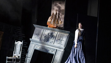 There is a chair and a fireplace that is falling. Above the fire place is a ship. Dido stands next to the fire place in a black dress.