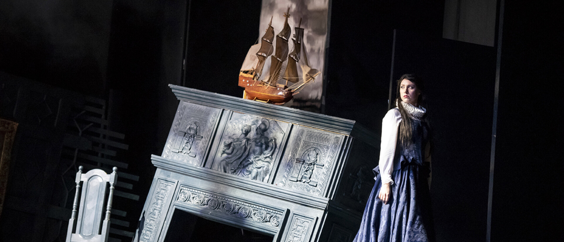 There is a chair and a fireplace that is falling. Above the fire place is a ship. Dido stands next to the fire place in a black dress.