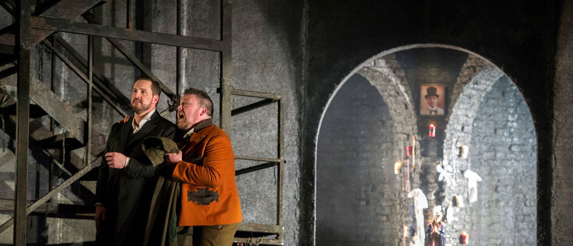Don Giovanni and Leporello are standing stage right next to a staircase. Leporello is holding his coat and Don Giovanni.