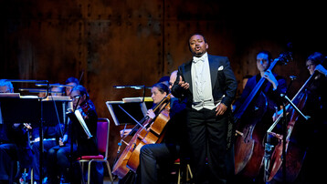 A man stands in full black tie singing in front of the cellos and double basses of the orchestra