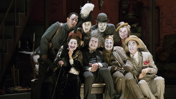 A group is huddle round Simone. Their faces are painted in a grotesque style and they are all looking out into the distance